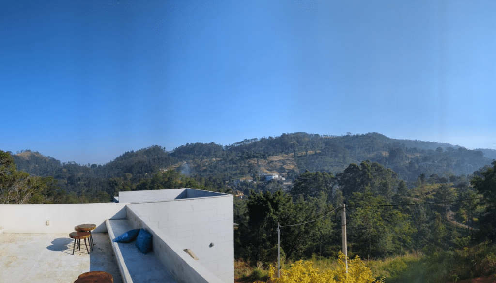 The Ultimate Guide to Redefining Remote Work in Sri Lanka with the Island Nomad Pass 4 "A scenic view from a modern white rooftop terrace in Sri Lanka, featuring wooden stools and blue cushions, overlooking lush green rolling hills and mountains under a clear blue sky."