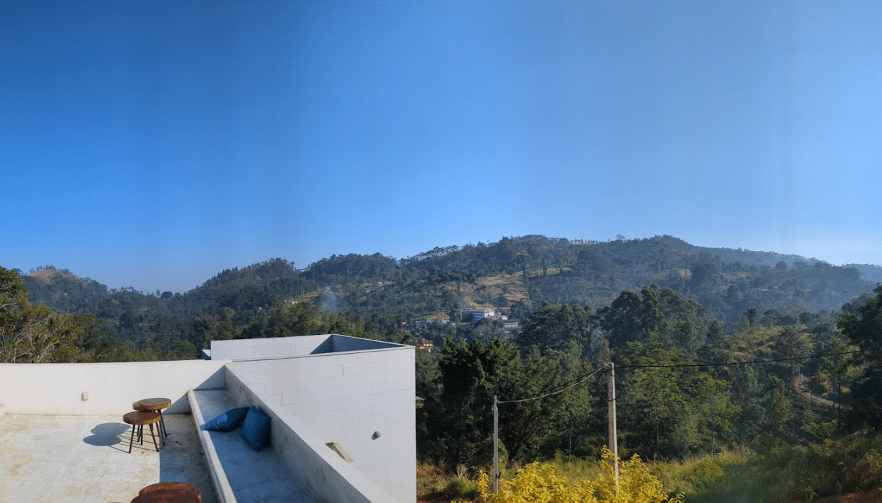 "A scenic view from a modern white rooftop terrace in Sri Lanka, featuring wooden stools and blue cushions, overlooking lush green rolling hills and mountains under a clear blue sky."